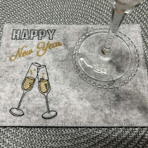 Puede incluir: Un posavasos de fieltro gris con texto bordado "HAPPY New Year" y dos copas de champ&aacute;n. Una copa de champ&aacute;n transparente descansa sobre el posavasos. El posavasos est&aacute; sobre un mantel individual gris tejido.