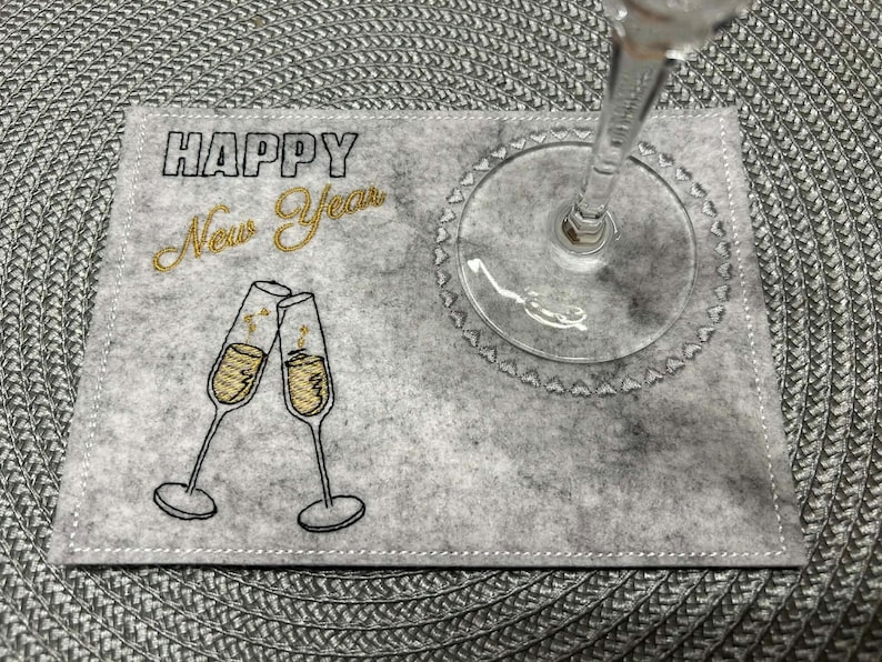 Puede incluir: Un posavasos de fieltro gris con texto bordado "HAPPY New Year" y dos copas de champ&aacute;n. Una copa de champ&aacute;n transparente descansa sobre el posavasos. El posavasos est&aacute; sobre un mantel individual gris tejido.