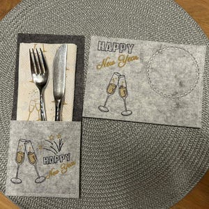Puede incluir: Un juego de manteles individuales de fieltro gris con un bolsillo para cubiertos y un posavasos separado. El mantel individual y el posavasos est&aacute;n decorados con copas de champ&aacute;n y las palabras "Happy New Year" en letras doradas.