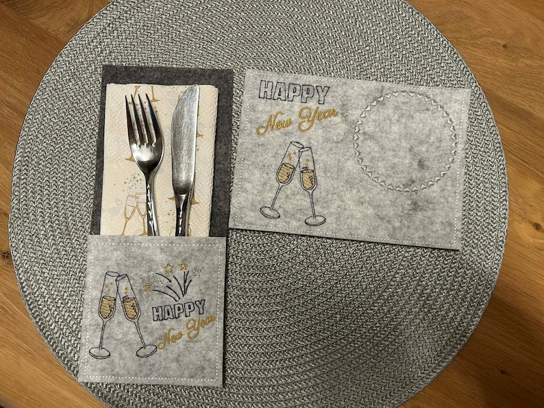 Puede incluir: Un juego de manteles individuales de fieltro gris con un bolsillo para cubiertos y un posavasos separado. El mantel individual y el posavasos est&aacute;n decorados con copas de champ&aacute;n y las palabras "Happy New Year" en letras doradas.