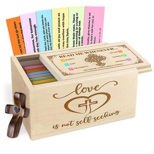 May include: Wooden box with a hinged lid, containing colorful cards with Bible verses. The box is engraved with a cross inside a heart and the words "love is not self seeking." A wooden cross is on the side.