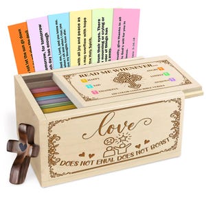 Op de afbeelding: Een houten kist met een scharnierend deksel, gegraveerd met het woord "Love" en bijbelverzen. De kist bevat kleurrijke kaarten met bijbelverzen. Een houten kruis is aan de zijkant bevestigd. De tekst op de kist luidt "Does Not Envy. Does Not Boast."