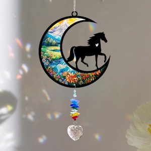 May include: A decorative suncatcher with a black horse silhouette within a crescent moon. The moon displays a colourful landscape with mountains, flowers, and a river. A rainbow crystal strand and a heart-shaped crystal hang below.