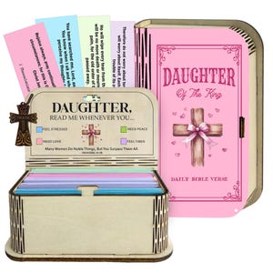 May include: A wooden box with a lid, containing colourful cards with Bible verses. The box is engraved with the words "Daughter, Read Me Whenever You..." and has a cross-shaped charm. A pink book with the words "Daughter of The King" is also visible.