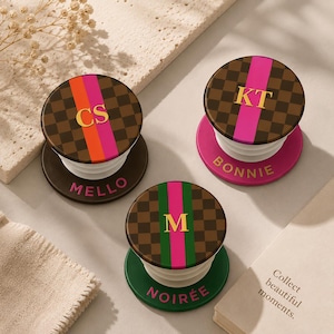May include: Three personalized phone grips with brown checkered tops, featuring initials and names. Each grip has a different color accent: orange and pink, pink, and green. The bases are dark brown, pink, and green, respectively. The text "MELLO", "BONNIE", and "NOIRÉE" are visible.