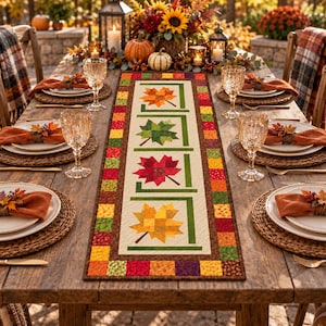 May include: A rectangular fabric table runner featuring a central white panel with four quilted maple leaf motifs in orange, green, red, and yellow, surrounded by a border of multicoloured patchwork squares in autumn tones.