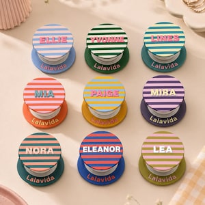 May include: Nine circular phone grips arranged in a grid, each featuring horizontal stripes in various color combinations and a personalized name printed in the center. Each base is printed with the brand name Lalavida in a small font.