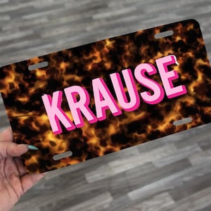 May include: A license plate with a fiery, tortoiseshell-like pattern. The word "KRAUSE" is displayed in large, pink letters with a white outline. The plate has four mounting holes.