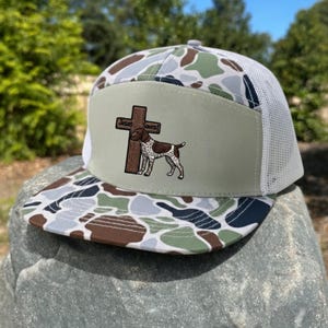 May include: A camouflage-patterned trucker hat with a white mesh back. The front panel features a brown cross and a dog illustration, with the words "Jesus Saves" above the dog.