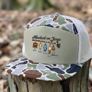 May include: A camouflage trucker hat with a white mesh back and a curved brim. The front panel features the text "Hooked on Jesus" with religious symbols and the Bible verse Mark 1:17.