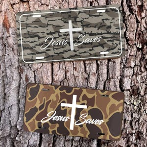 May include: Two license plates with camouflage patterns. One plate has a gray and brown camouflage design, the other has a brown and tan camouflage design. Both plates feature a white cross and the text "Jesus Saves".
