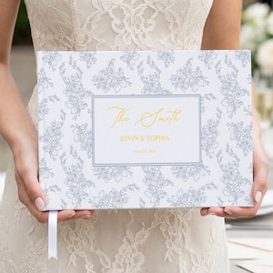 Peut inclure: Un livre d'or blanc avec un motif floral gris et bleu est tenu. Le livre présente le texte "The Smith" en or, ainsi que les noms Kevin & Sophia et une date. Un signet en ruban blanc est attaché.