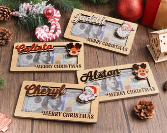 Könnte beinhalten: Weihnachtliche Holzrahmen mit Namen, jeweils mit einem 100-Dollar-Schein. Die Rahmen weisen dekorative Elemente wie den Weihnachtsmann und Rentiere auf, mit der Aufschrift "MERRY CHRISTMAS". Die Namen "Judith", "Calista", "Cheryl" und "Alston" sind zu sehen.