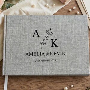 Könnte beinhalten: Hellgraues Gästebuch zur Hochzeit mit Leinenstruktur. Auf dem Cover stehen die Namen "Amelia & Kevin" und das Datum "25. Februar 2026" in Schwarz. Die Buchstaben "A" und "K" sind mit einem floralen Design versehen.
