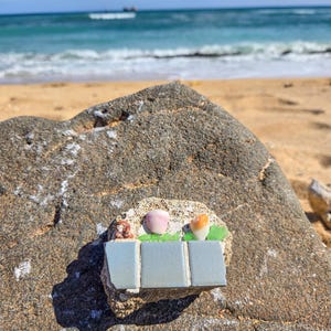 May include: A mosaic art piece featuring a light blue rectangular design with three sections, adorned with seashells and sea glass. The artwork is set on a large, textured rock, with a beach and ocean backdrop.