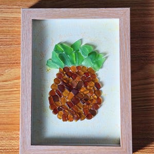 May include: Framed artwork depicting a mosaic pineapple. The pineapple is made from amber and brown glass pieces, with green glass leaves. The artwork is set against a light background and framed in a light wood frame.