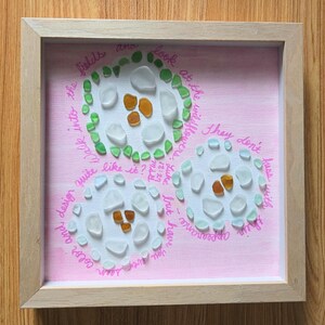 May include: Framed artwork with three floral designs made of sea glass on a pink background. The sea glass is white, green, and amber. Handwritten text in pink surrounds the designs, with a light wood frame.