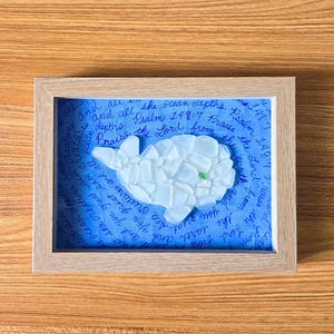May include: Framed artwork with a white whale mosaic on a blue background, featuring handwritten text. The whale is made of small, irregular white pieces. The frame is light-coloured wood. The text includes the words "Praise the Lord".