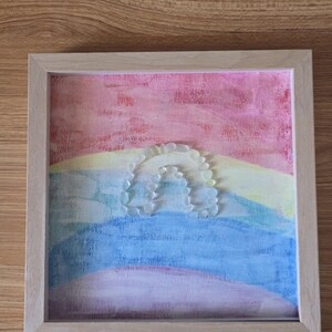 Puede incluir: Obra de arte enmarcada con un diseño de arcoíris. El fondo está pintado con tonos pastel de rosa, amarillo, azul y morado. Un arcoíris está formado por piedras claras y redondeadas. El marco es de madera clara.