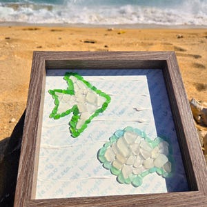 May include: Framed artwork with a fish and cloud design crafted from sea glass. The fish is outlined in green, and the cloud has a green and blue outline. The background has faint text. The frame is a light brown colour.