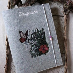 Puede incluir: Un portadocumentos de fieltro gris con la palabra "Zeugnisse" bordada en blanco. El portadocumentos presenta un gato bordado con una mariposa y una flor roja. Un cordón blanco con cuentas rosas asegura el portadocumentos.