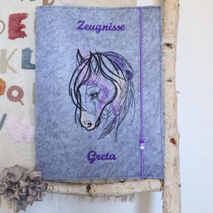 Op de afbeelding: Een grijze vilten hoes met een paarse paardenhoofdborduur, het woord "Zeugnisse" en de naam "Greta" in paars schrift. Een paars elastisch koord met een kralensluiting bevindt zich aan de rechterkant. De hoes hangt aan een houten frame.