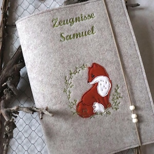 以下が含まれることがあります： 緑色の刺繍で「Zeugnisse Samuel」と書かれた、薄い灰色のフェルトカバー。赤と白のキツネが下に刺繍され、緑のリースで囲まれています。白いビーズが付いたベージュのゴム紐がカバーを固定しています。