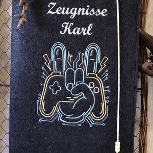 May include: A dark gray felt document folder featuring the embroidered text Zeugnisse Karl and a graphic of a hand making a peace sign over a video game controller, accented with light blue and yellow circuit board lines and a white elastic band.
