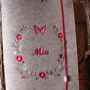May include: A light brown felt notebook cover featuring an embroidered floral wreath in shades of pink, purple, and green. A pink butterfly is stitched at the top of the wreath, and the name Mia is embroidered in pink in the center. A pink elastic band with three small beads secures the cover.