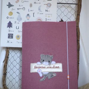 Puede incluir: Una carpeta de documentos de fieltro rosa polvoriento con una aplicación de gato gris que sostiene un parche rectangular blanco con el texto bordado Zeugnisse von Lena. La carpeta se asegura con una banda elástica blanca con dos cuentas.