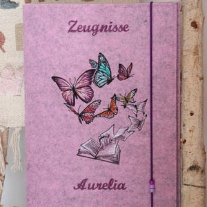 May include: A pink felt journal cover with embroidered butterflies in various colors. The word "Zeugnisse" is embroidered at the top, and "Aurelia" at the bottom. A purple elastic band secures the journal.