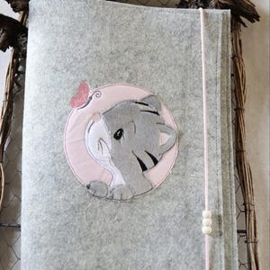 Puede incluir: Una cubierta de libro de fieltro gris con un diseño bordado de un cachorro de tigre gris dentro de un círculo rosa, con una mariposa rosa. Una cuerda rosa con tres cuentas blancas asegura la cubierta. La cubierta está sobre un fondo rústico de mimbre.