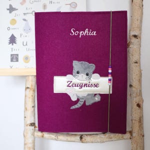 May include: A deep purple felt cover embroidered with the name "Sophia" in white. A grey cat illustration holds a banner with the word "Zeugnisse". A string with beads is attached. An alphabet poster is in the background.