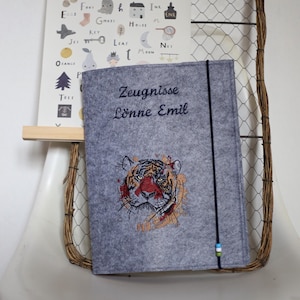May include: A gray felt binder with the text "Zeugnisse Lönne Emil" and an embroidered tiger face. A poster with the alphabet and illustrations hangs behind the binder. The binder is held closed with a black elastic band.