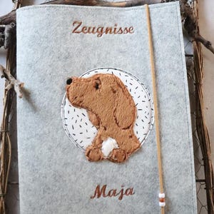 Puede incluir: Una carpeta de documentos de fieltro gris claro con el perfil de un perro marrón bordado sobre un fondo circular blanco con rayas negras. El texto Zeugnisse está bordado en marrón sobre el perro y el nombre Maja está bordado debajo.