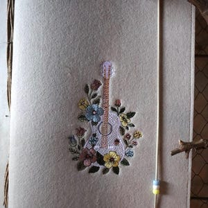May include: A beige felt notebook cover featuring a central embroidered design of an acoustic guitar surrounded by colorful flowers in shades of blue, yellow, pink, and green. A vertical elastic band with three small beads secures the cover.