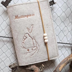 Könnte beinhalten: Eine hellgraue Filzhülle mit dem in braunem Garn gestickten Schriftzug Mutterpass. Das Design zeigt die braune Stickerei einer Hand, die einen Babyfuß hält, mit einem kleinen Herz darüber. Ein goldenes Gummiband mit drei Holzperlen.