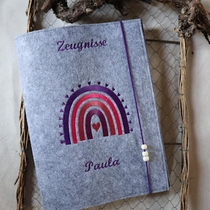 May include: A gray felt document holder with a rainbow design in shades of purple and red. The holder has the word "Zeugnisse" at the top and the name "Paula" at the bottom, both in purple embroidery.