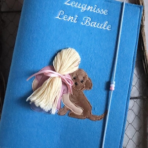 Puede incluir: Una carpeta azul de fieltro con texto bordado en blanco que dice "Zeugnisse Leni Baule". La portada presenta una aplicación de una niña abrazando a un perro marrón, con un lazo rosa. Un cordón elástico azul claro asegura la carpeta.