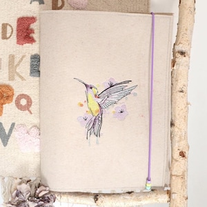 Può includere: Una copertina per diario in tessuto beige con un ricamo di colibrì in giallo, viola e verde, circondato da fiori viola. Un elastico viola assicura la copertina. Presentato su un ramo di betulla.