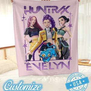 May include: A pink blanket featuring a graphic of three stylized female figures and a blue cartoon cat. The blanket has the text "HUNTRAX" and "EVELYN" in purple. The blanket is held up by a person.