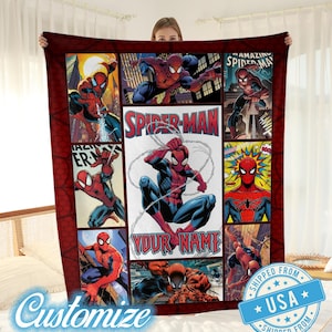 Op de afbeelding: Een rood-blauwe Spider-Man deken met een collage van stripboekpanelen. De deken heeft de tekst "SPIDER-MAN" en "YOUR NAME". De deken wordt door een persoon vastgehouden.