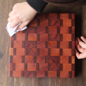 May include: A square, end-grain butcher block cutting board with a checkerboard pattern of reddish-brown and dark brown wood squares. The board is being wiped with a white cloth. The wood grain is visible.