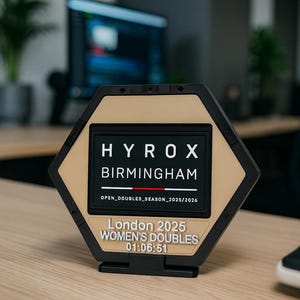 May include: A hexagonal trophy with a black frame and a tan center plate. The text reads HYROX BIRMINGHAM, OPEN_DOUBLES_SEASON_2025/2026, London 2025, WOMEN'S DOUBLES, and 01:06:51. The trophy features a red horizontal line below the word BIRMINGHAM.