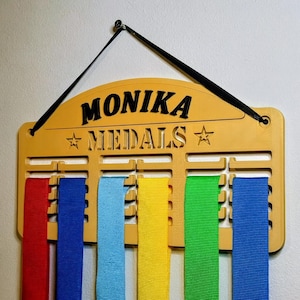 May include: A wooden medal display with the name "MONIKA" and the word "MEDALS" in black lettering. The display has slots for ribbons in red, blue, light blue, yellow, green, and dark blue. A black cord is attached for hanging.