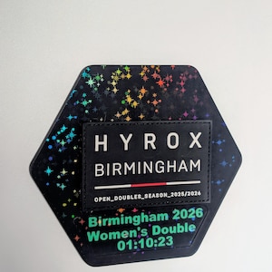 May include: A hexagonal black patch featuring a holographic star pattern. A rectangular black label is centered with the white text HYROX BIRMINGHAM, OPEN_DOUBLES_SEASON_2025/2026, and the green text Birmingham 2026 Women's Double 01:10:23.