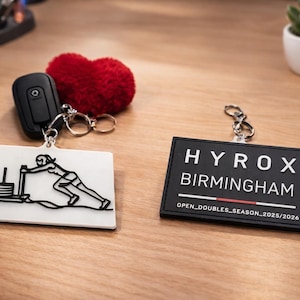 May include: Two keychains with silver rings and a car key. One keychain is white with a black graphic of a person pushing a weight. The other is black with the text "HYROX BIRMINGHAM" in white. A red heart-shaped item is also present.
