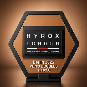 Puede incluir: Un trofeo hexagonal con marco negro y centro color bronce. La sección superior presenta una placa rectangular negra con texto blanco que dice HYROX LONDON y OPEN_DOUBLES_SEASON_2025/2026. Debajo, el texto blanco indica Berlin 2026 DOUBLES 1:18:56.