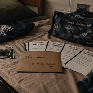 May include: A collection of white cards with black text including phrases like Open When You Need To Laugh, Open When You Want To Quit, Open When You Miss Home, and Open When You Need Encouragement, alongside a brown envelope labeled Open when you miss home.