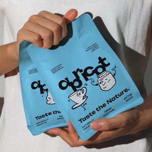 Puede incluir: Dos paquetes de café "Apricot" de color azul claro se sostienen frente a una camiseta blanca. Los paquetes tienen el texto "Taste the Nature" y "Fresh Mind Fuel". Los paquetes también tienen ilustraciones de dibujos animados.